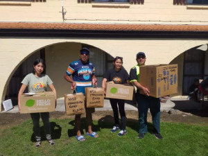 Four people hold boxes and bags marked as food. They are standing in front of a building.