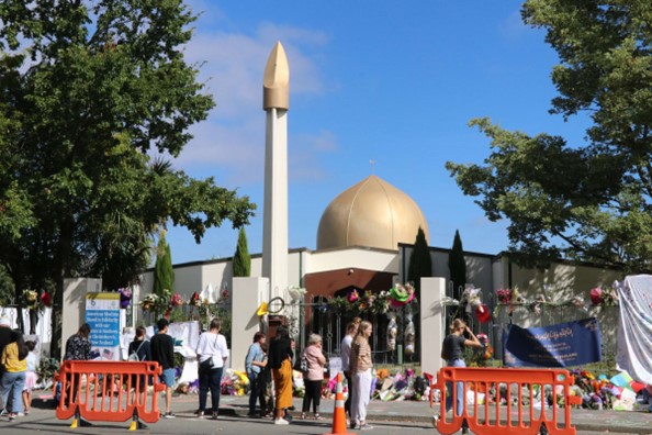 Al-Noor Mosque in the background with the fence in front covered with flowers, and people placing flowers in front of the Mosque.