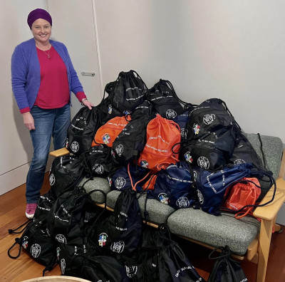 A woman stands next to a couch with a massive pile of bags on it. Bags are also laid across the flood. These are emergency grab bags.